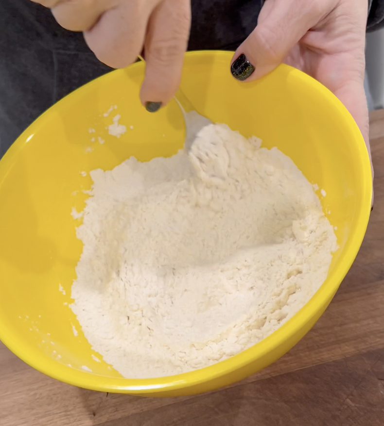 A person mixes flour in a bright yellow bowl with a fork on a wooden surface.