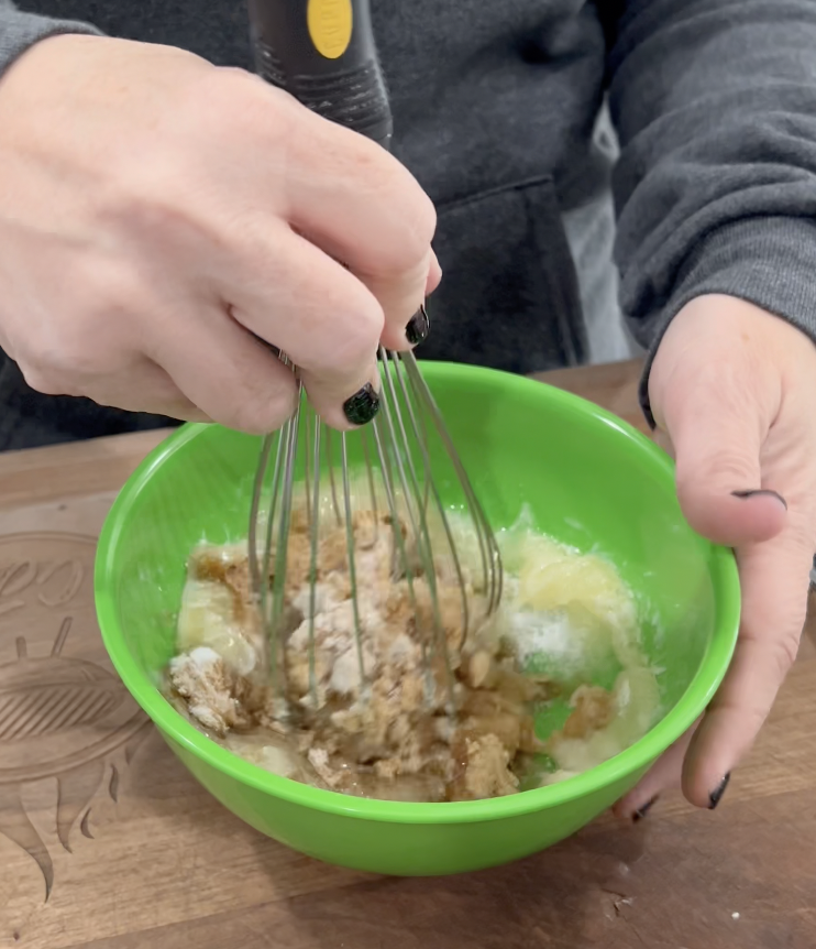 A person whisking brown and white ingredients in a bright green bowl on a wooden surface. The person is wearing a dark sweatshirt and has black nail polish.