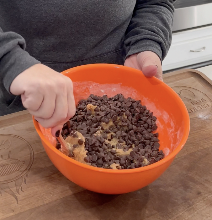 A person wearing a dark hoodie mixes cookie dough and chocolate chips in an orange bowl on a wooden cutting board in a kitchen.