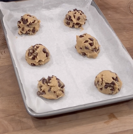 Six scoops of chocolate chip cookie dough are spaced on a parchment-lined baking sheet, ready to be baked, sitting on a wooden surface.