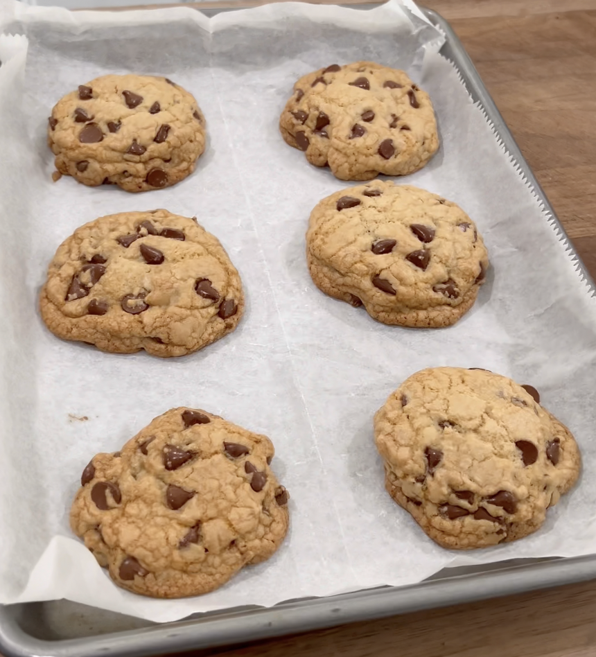 Six freshly baked chocolate chip cookies rest on a parchment-lined baking sheet atop a wooden surface.