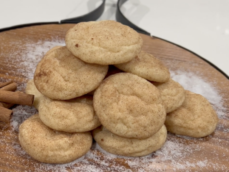 A stack of snickerdoodle cookies dusted with cinnamon sugar sits on a wooden board, next to cinnamon sticks and sprinkled flour.
