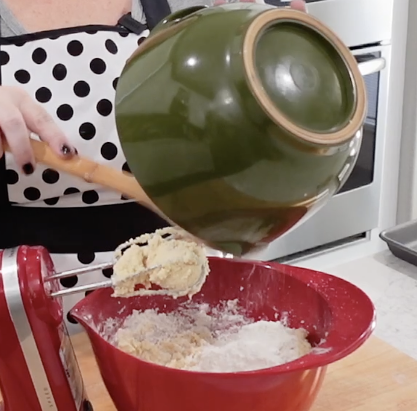 A person wearing a polka dot apron pours dough from a green bowl into a red mixing bowl using a wooden spoon. An electric mixer is nearby on the counter.