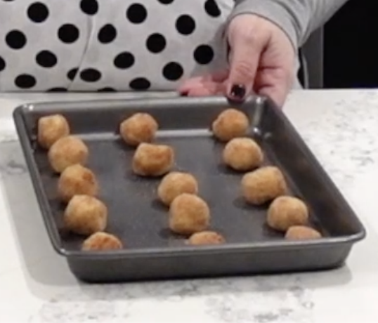 A person holds a baking pan with evenly spaced, round dough balls ready to be baked. The person wears a gray shirt and a white apron with black polka dots.