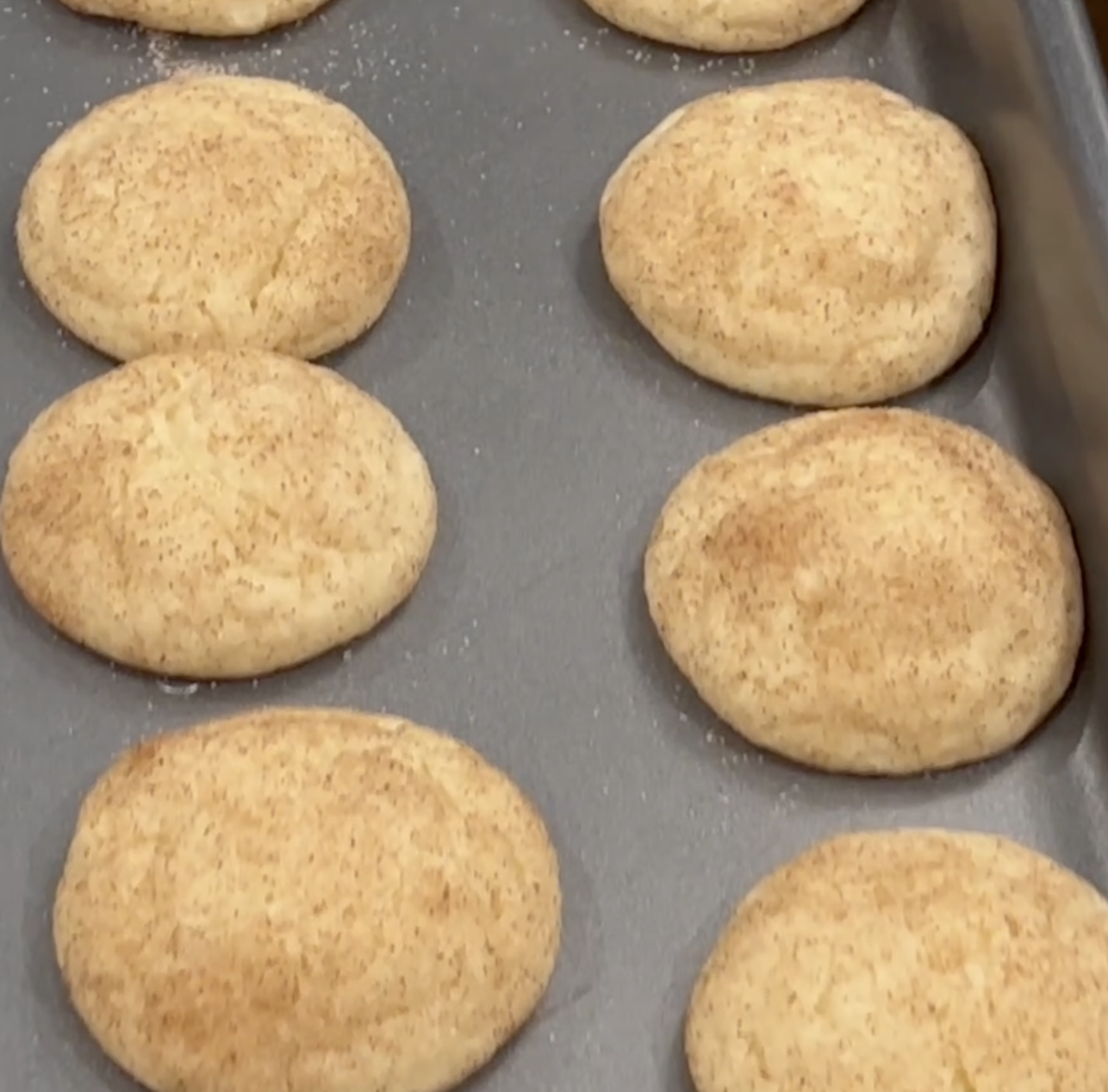 A baking tray with eight golden-brown snickerdoodle cookies, evenly spaced and freshly baked, showing a light dusting of cinnamon sugar on top.