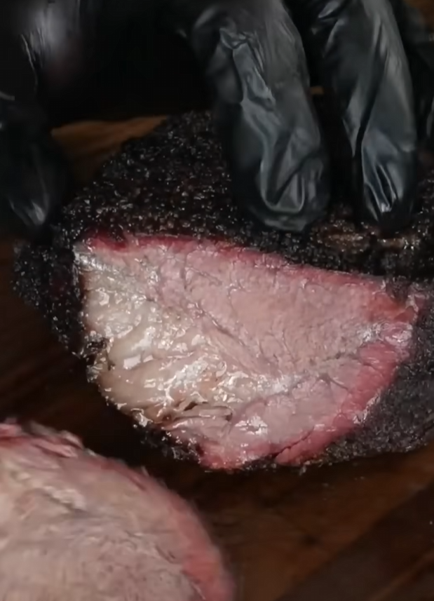 A gloved hand holds a sliced brisket with a dark bark and a visible pink smoke ring, showcasing a juicy, tender interior on a wooden cutting board.