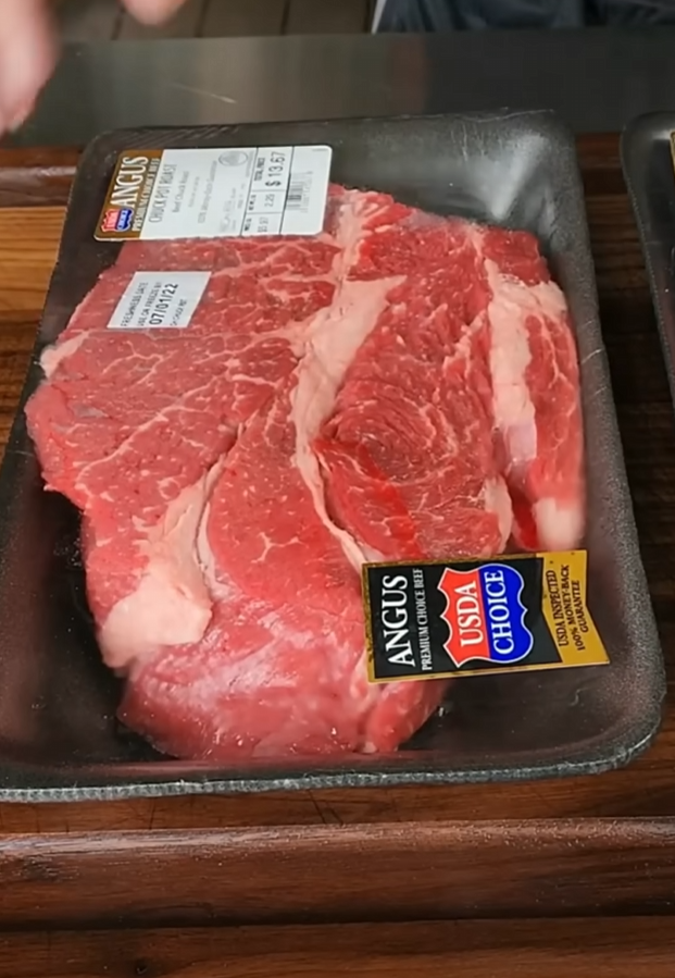 Packaged raw Angus beef steaks labeled USDA Choice sit on a black tray with stickers showing the cut and price, placed on a wooden surface.