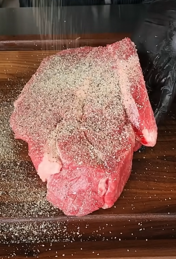 A gloved hand seasons a large raw steak with salt and pepper on a wooden cutting board, with seasoning visibly falling onto the meat.