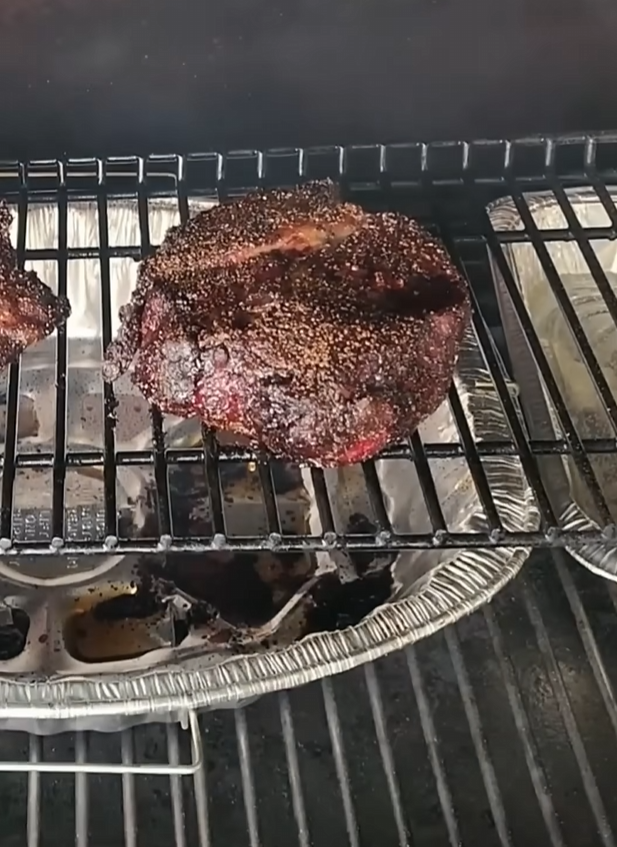 A thick, seasoned steak is being smoked on a grill over metal racks, with disposable aluminum trays underneath catching drippings.
