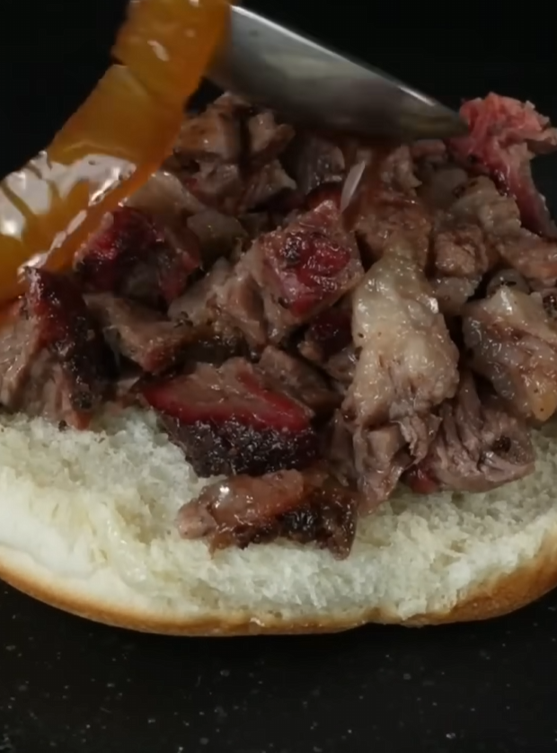 A close-up of chopped barbecued beef on a sandwich bun, with sauce being poured over the meat from a spoon.