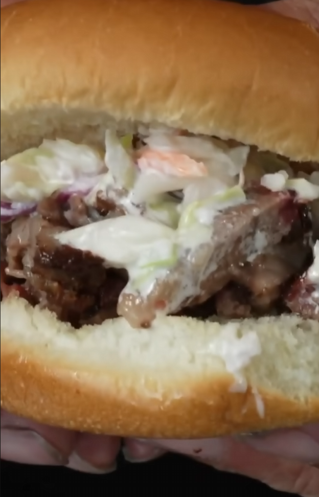 A close-up of a sandwich with sliced meat and coleslaw in a soft hamburger bun, held in someones hands.