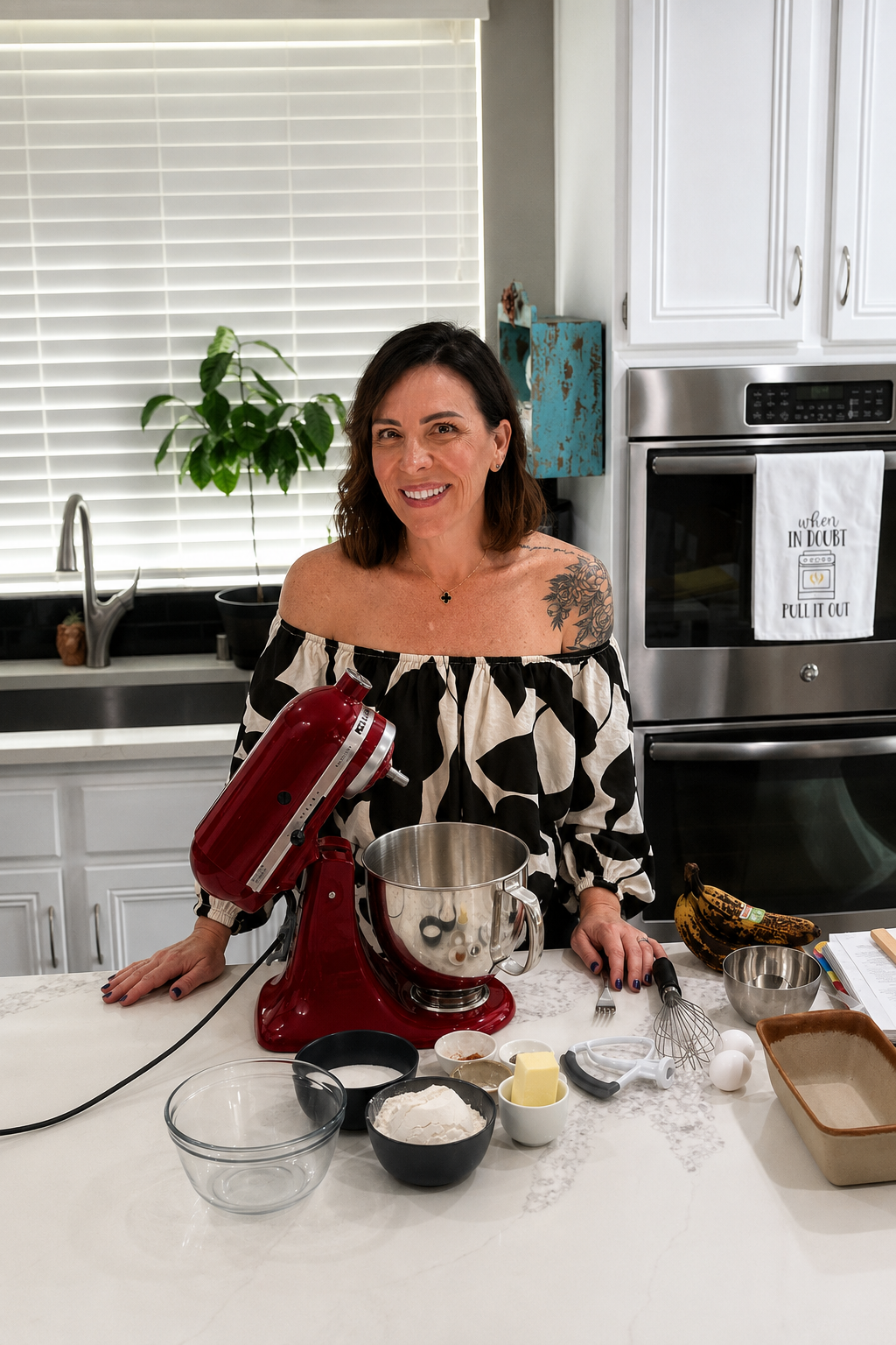 A woman stands in a kitchen behind a counter with baking ingredients, a mixer, and utensils. She smiles at the camera, wearing an off-shoulder dress. White cabinets and a potted plant are in the background.