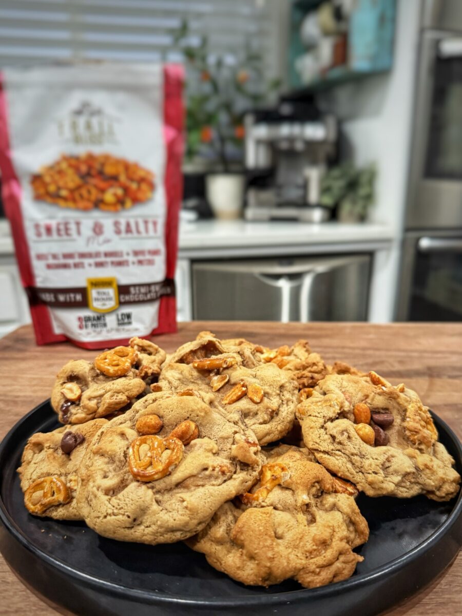 A black plate of chunky cookies with pretzels and chocolate chips sits on a wooden counter, with a bag of Sweet & Salty snack mix blurred in the background in a modern kitchen.