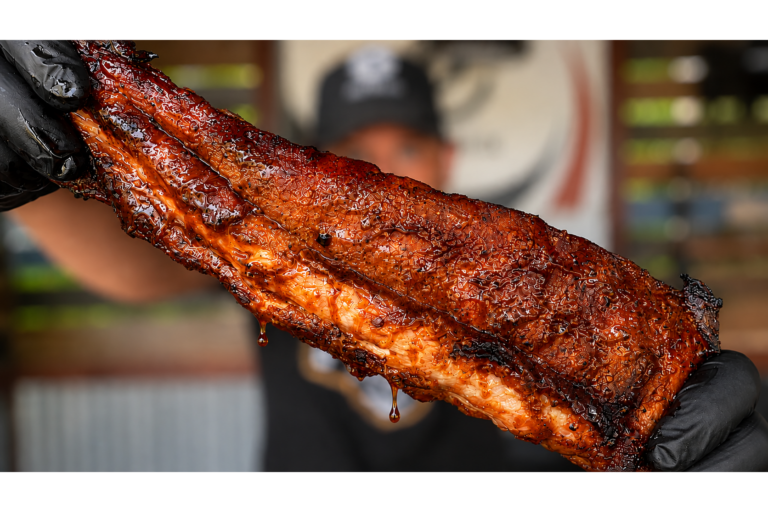 A person wearing black gloves holds up a large, juicy slab of barbecued ribs, glistening with sauce, in front of a blurred background.