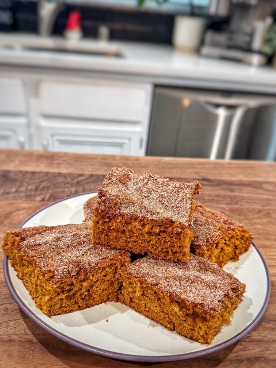 A plate of four square pumpkin cake bars topped with cinnamon sugar sits on a wooden counter in a bright kitchen.