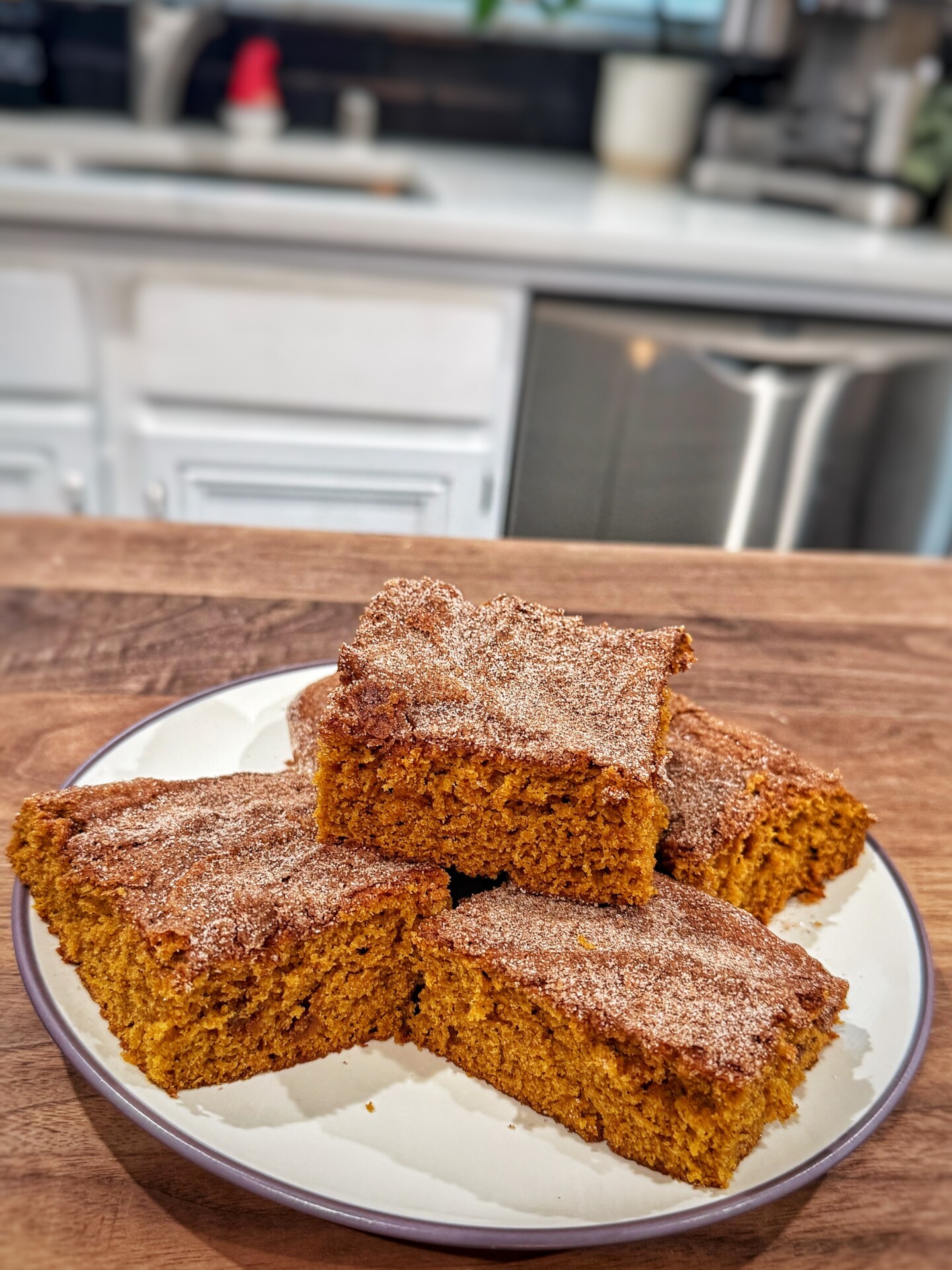 A plate of four square pumpkin cake bars topped with cinnamon sugar sits on a wooden counter in a bright kitchen.