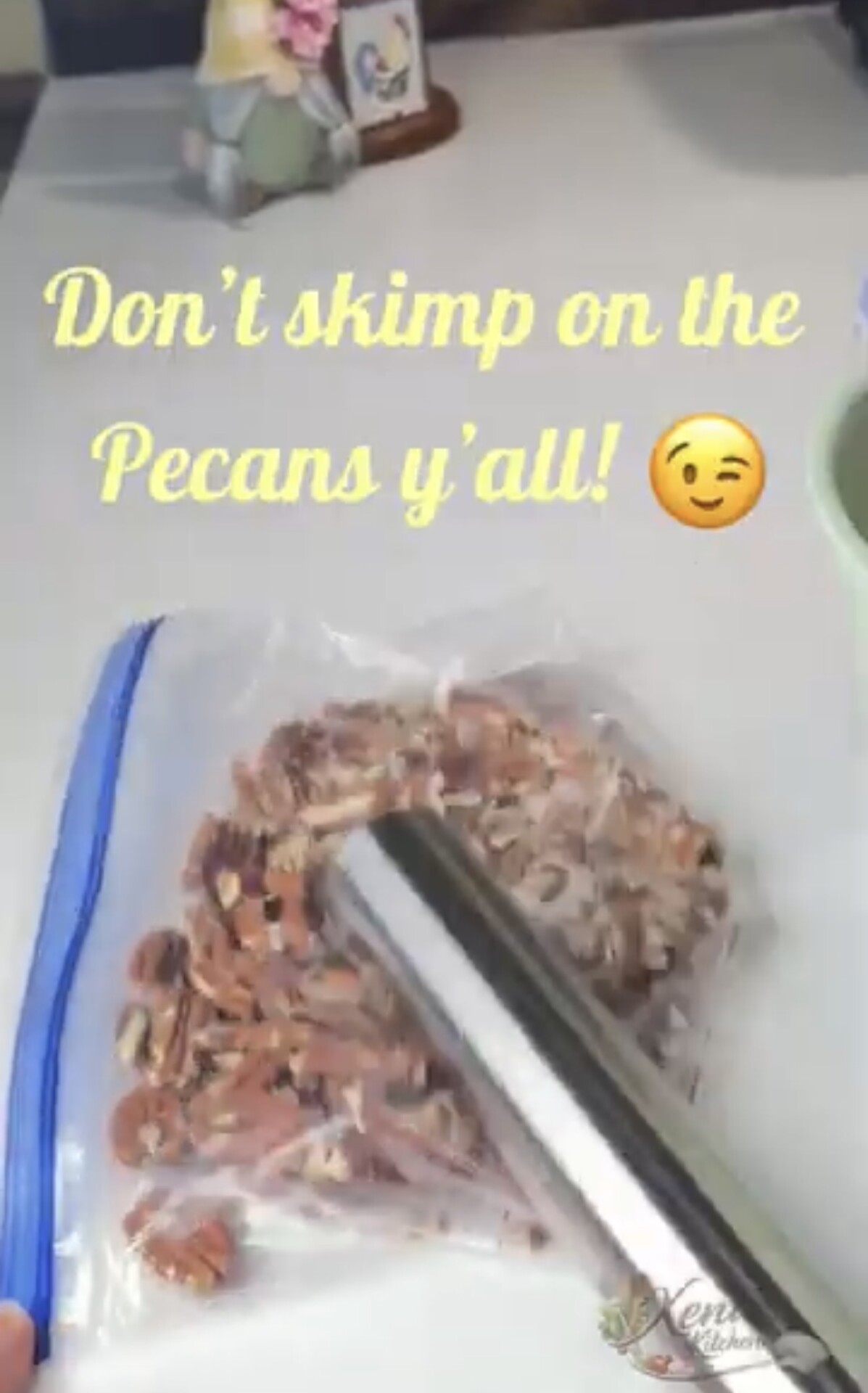 A hand crushes pecans in a zip-top bag with a metal rolling pin. Text reads, Dont skimp on the pecans yall! with a winking emoji. A green bowl and decor are in the background.
