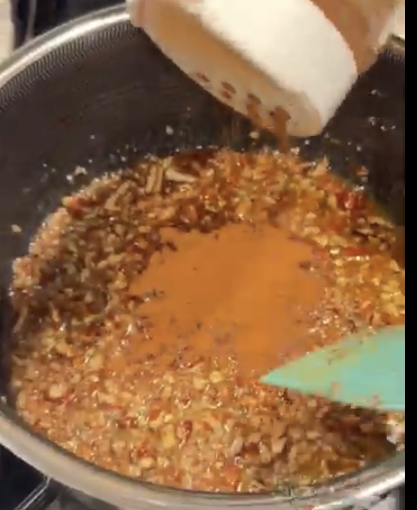 Ground cinnamon is being sprinkled from a container into a pan containing chopped nuts and spices, which are being stirred with a teal spatula.