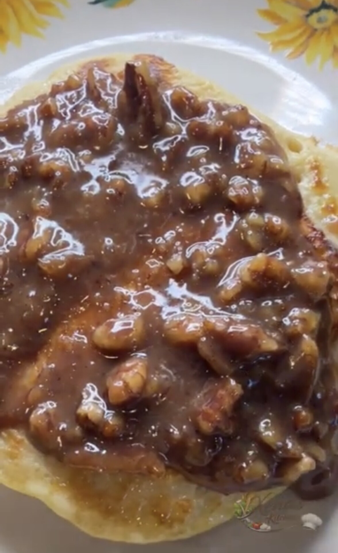 A close-up of a pancake topped with a glossy pecan and caramel sauce is shown on a plate with a sunflower pattern along the rim.