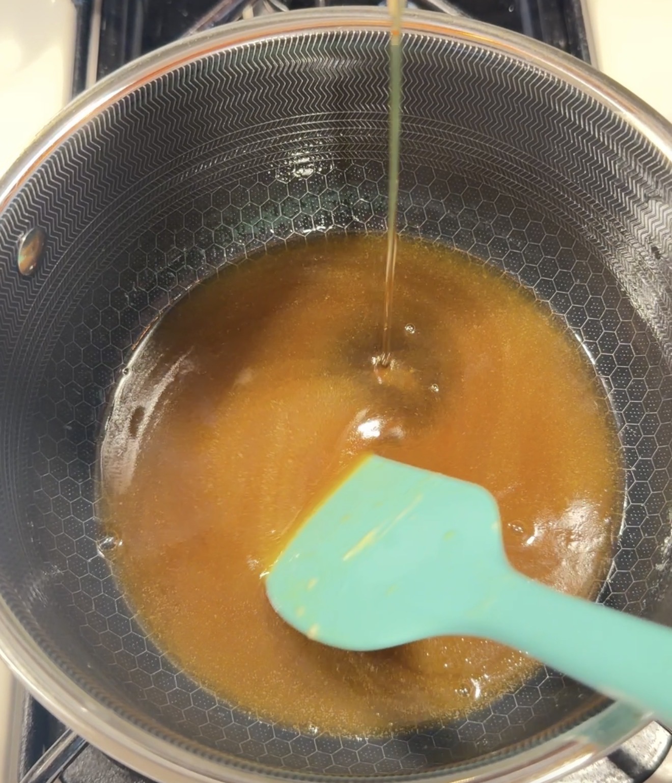 A blue spatula stirs a thick, golden-brown liquid in a saucepan with a honeycomb pattern, while more liquid is poured in from above.