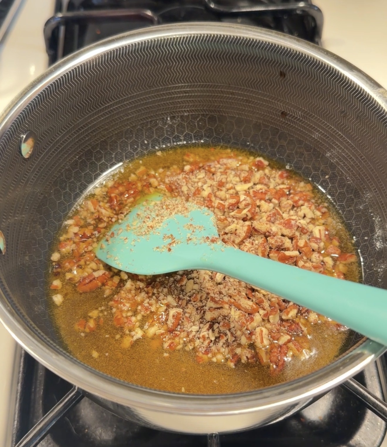 A saucepan on a stove contains melted butter and chopped pecans, being stirred with a teal spatula.