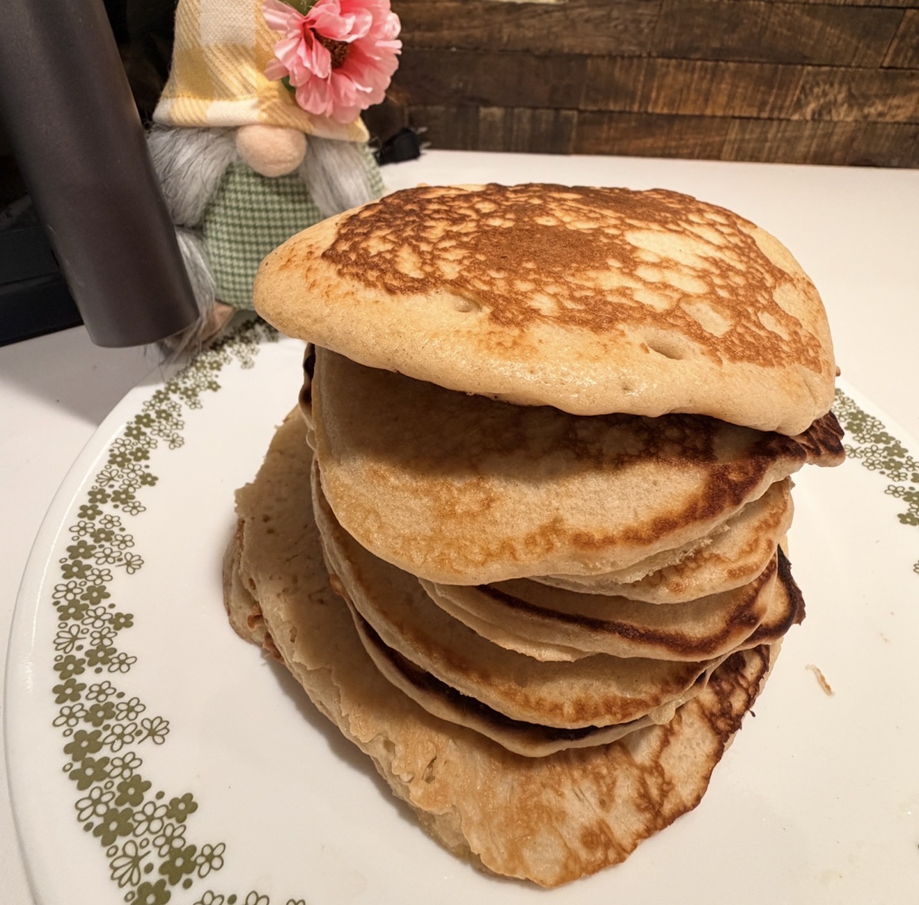 A stack of golden pancakes sits on a patterned plate, with a decorative gnome wearing a pink flower and green outfit in the background on a wooden surface.