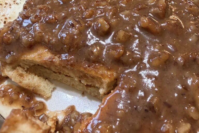 A close-up of pancakes covered in a thick, chunky brown sauce with visible pieces of fruit, possibly bananas. A wedge has been cut out, revealing the soft inside of the pancakes.