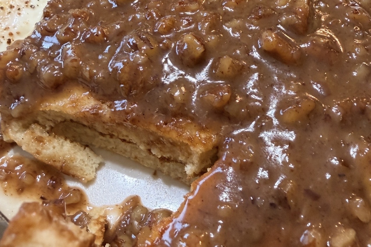 A close-up of pancakes covered in a thick, chunky brown sauce with visible pieces of fruit, possibly bananas. A wedge has been cut out, revealing the soft inside of the pancakes.