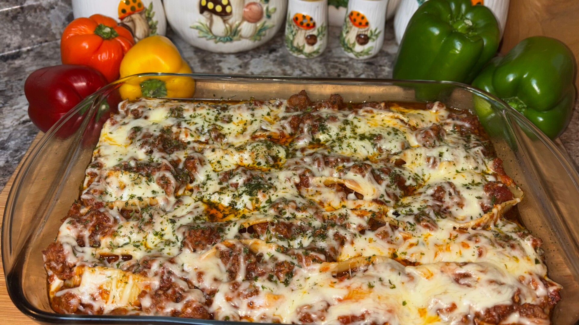 A glass baking dish filled with baked lasagna topped with melted cheese and herbs, with colorful bell peppers and kitchen decor in the background.