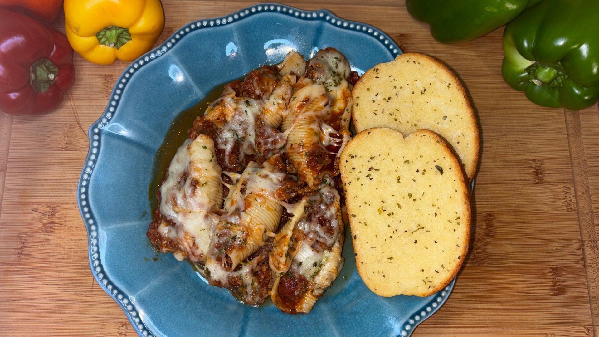 Stuffed pasta shells topped with melted cheese and tomato sauce on a blue plate, served with two slices of garlic bread. Bell peppers in red, yellow, and green are placed around the wooden surface.