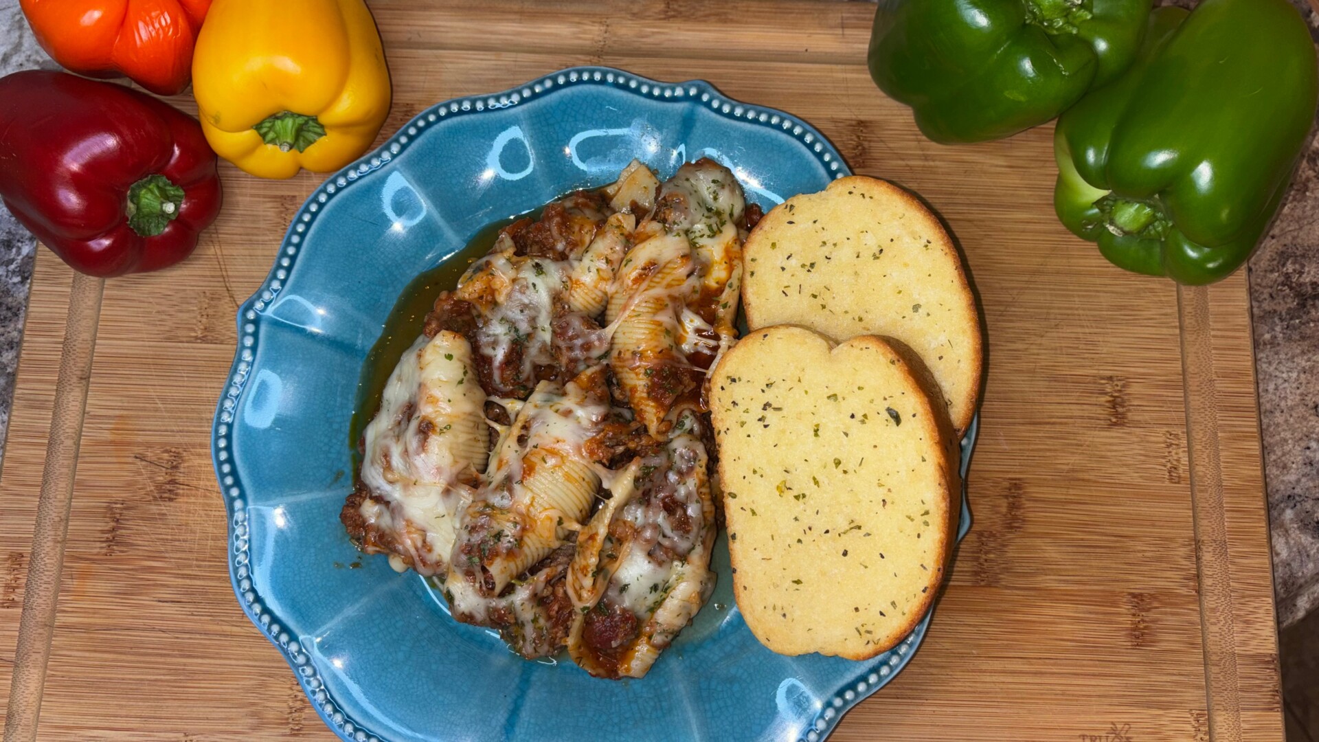 Stuffed pasta shells with cheese and sauce on a blue plate, served with two slices of garlic bread. Red, yellow, and green bell peppers are beside the plate on a wooden surface.