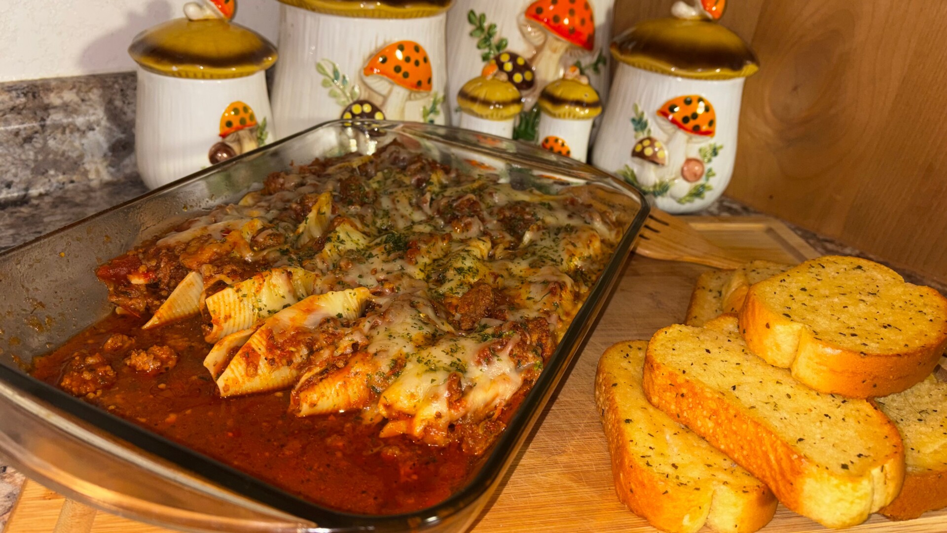 A glass baking dish of stuffed pasta shells with meat sauce and melted cheese sits on a wooden board next to slices of garlic bread. Mushroom-themed ceramic canisters are in the background.