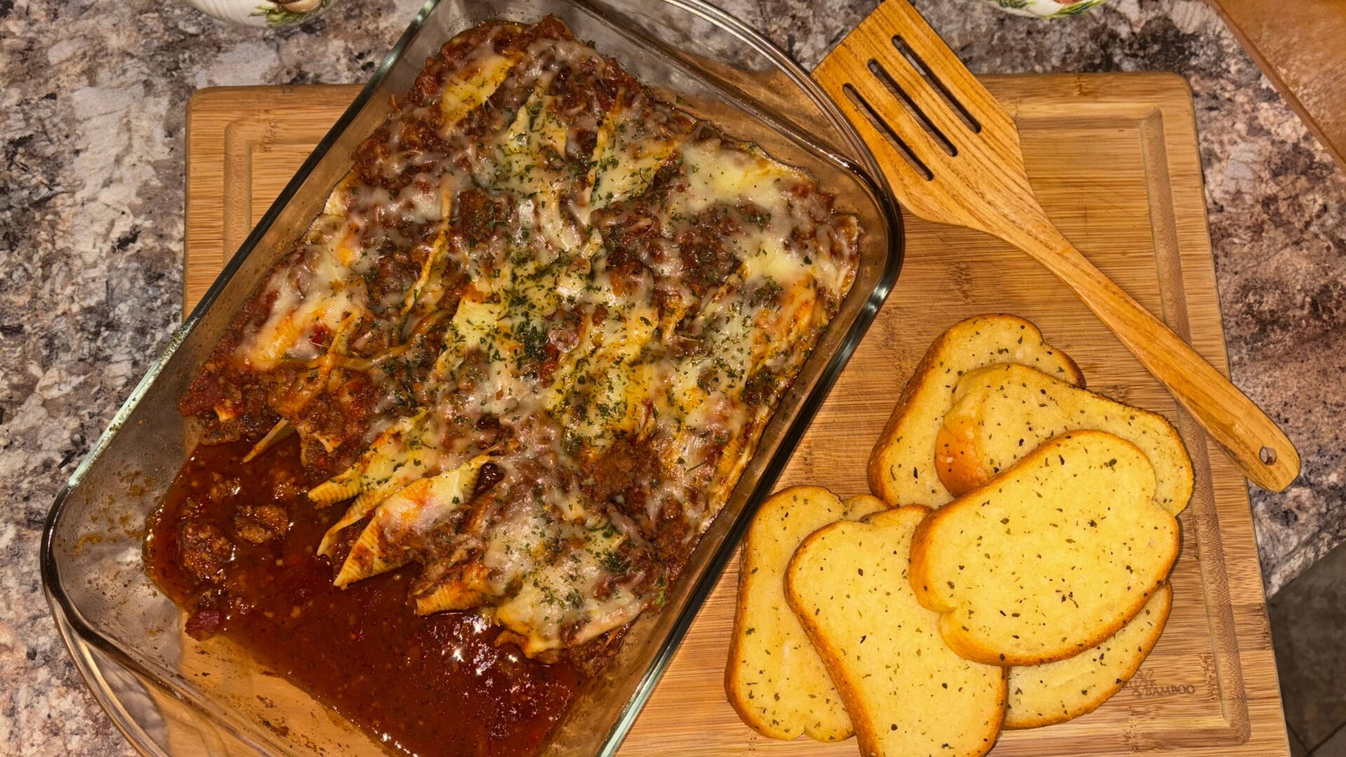 A glass dish of baked pasta with meat sauce and melted cheese sits on a wooden board next to a stack of toasted garlic bread slices and a wooden spatula.