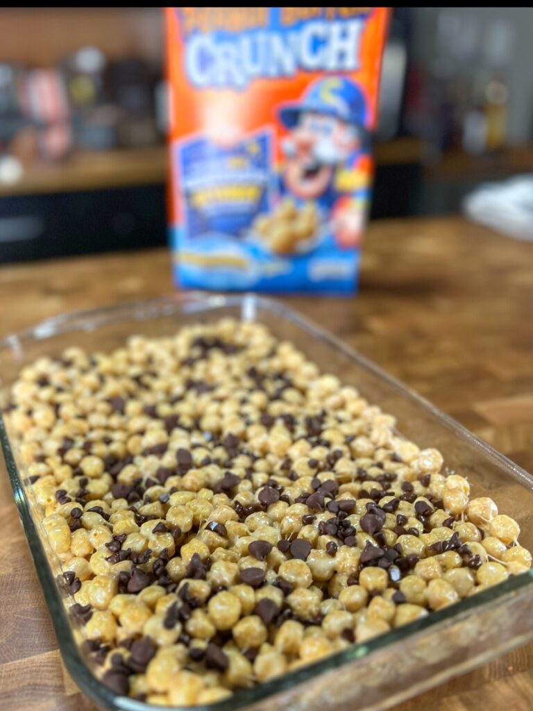 A glass baking dish filled with a dessert made of round cereal pieces and chocolate chips sits on a wooden counter, with a colorful Capn Crunch cereal box in the background.