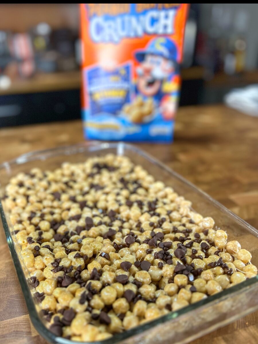 A glass baking dish filled with a dessert made of round cereal pieces and chocolate chips sits on a wooden counter, with a colorful Capn Crunch cereal box in the background.