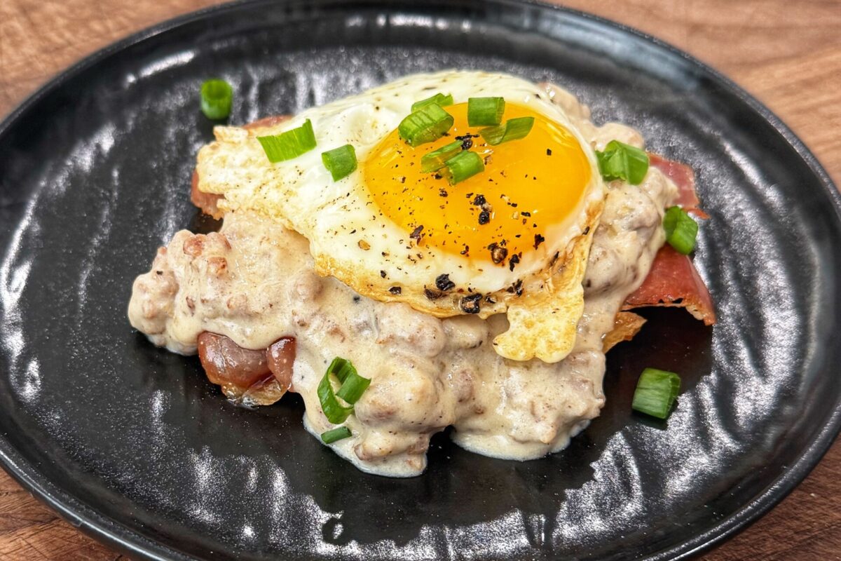 A black plate with sausage gravy over toast, topped with a sunny-side-up egg and garnished with chopped green onions.