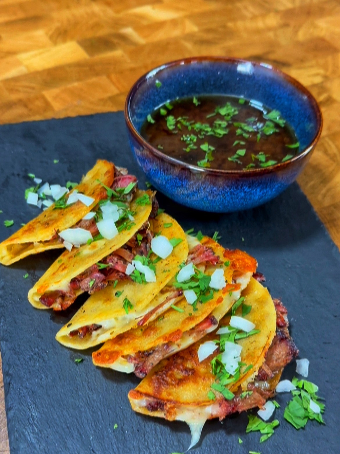 Four birria tacos filled with meat, topped with chopped onions and cilantro, are arranged on a slate with a bowl of dark consommé garnished with herbs on the side.