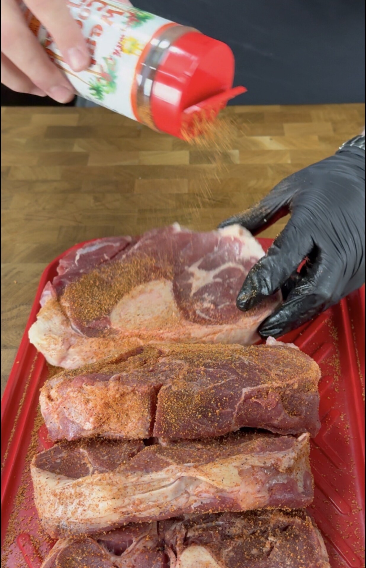 A person sprinkles seasoning onto several raw steaks on a red tray. One hand holds the seasoning container, while the other, wearing a black glove, arranges the meat on a wooden surface.