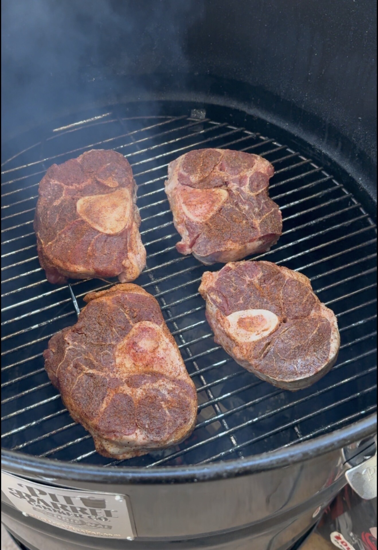 Four seasoned cuts of meat with visible bones are being smoked on a grill inside a black smoker, with smoke rising around them.