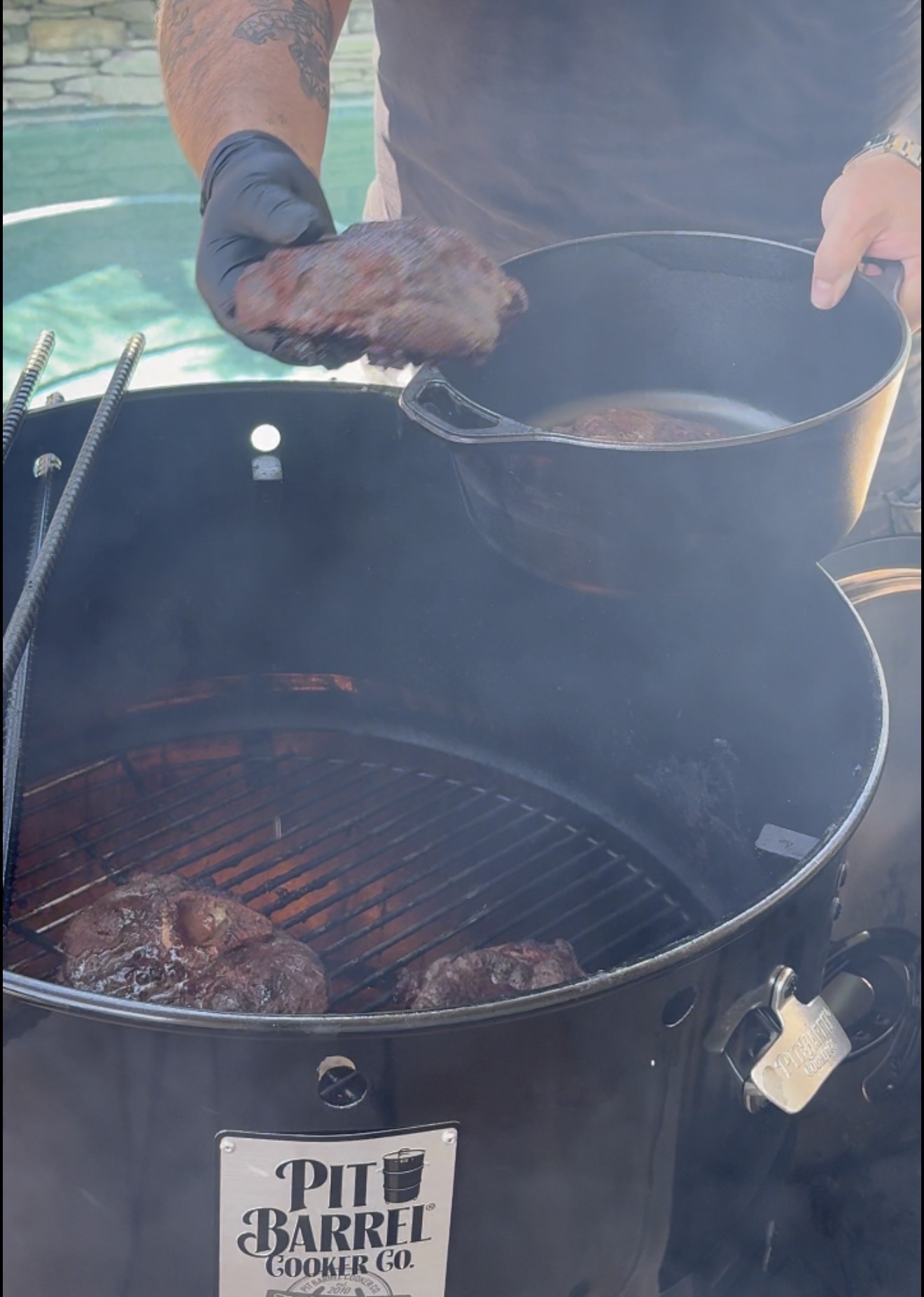 A person wearing black gloves lifts cooked meat from a grill smoker into a black pot. Smoke rises around the Pit Barrel Cooker, and another piece of meat remains on the grill.