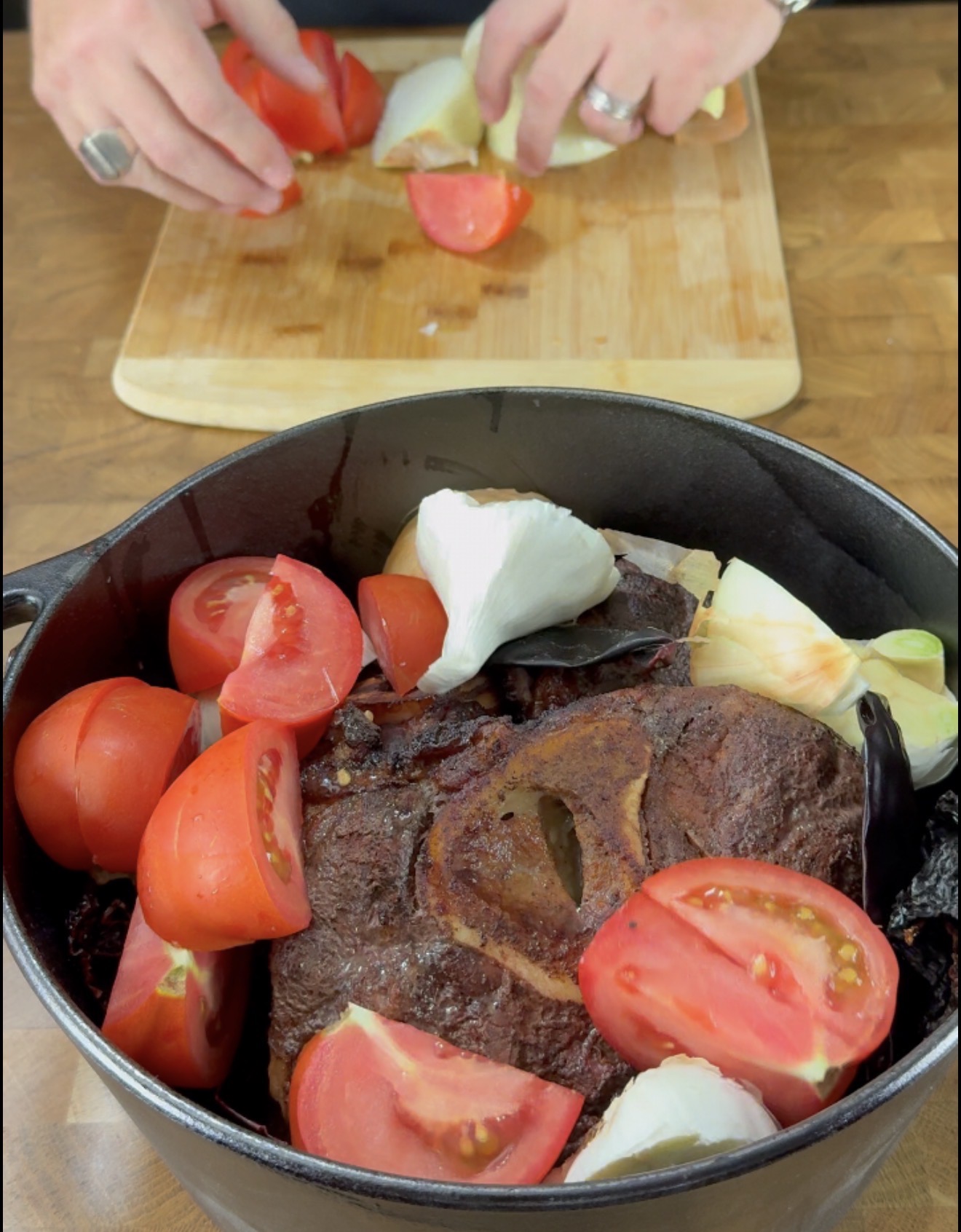 A black pot with a browned meat shank, quartered tomatoes, onions, and chilies. In the background, hands chop tomatoes on a wooden cutting board.