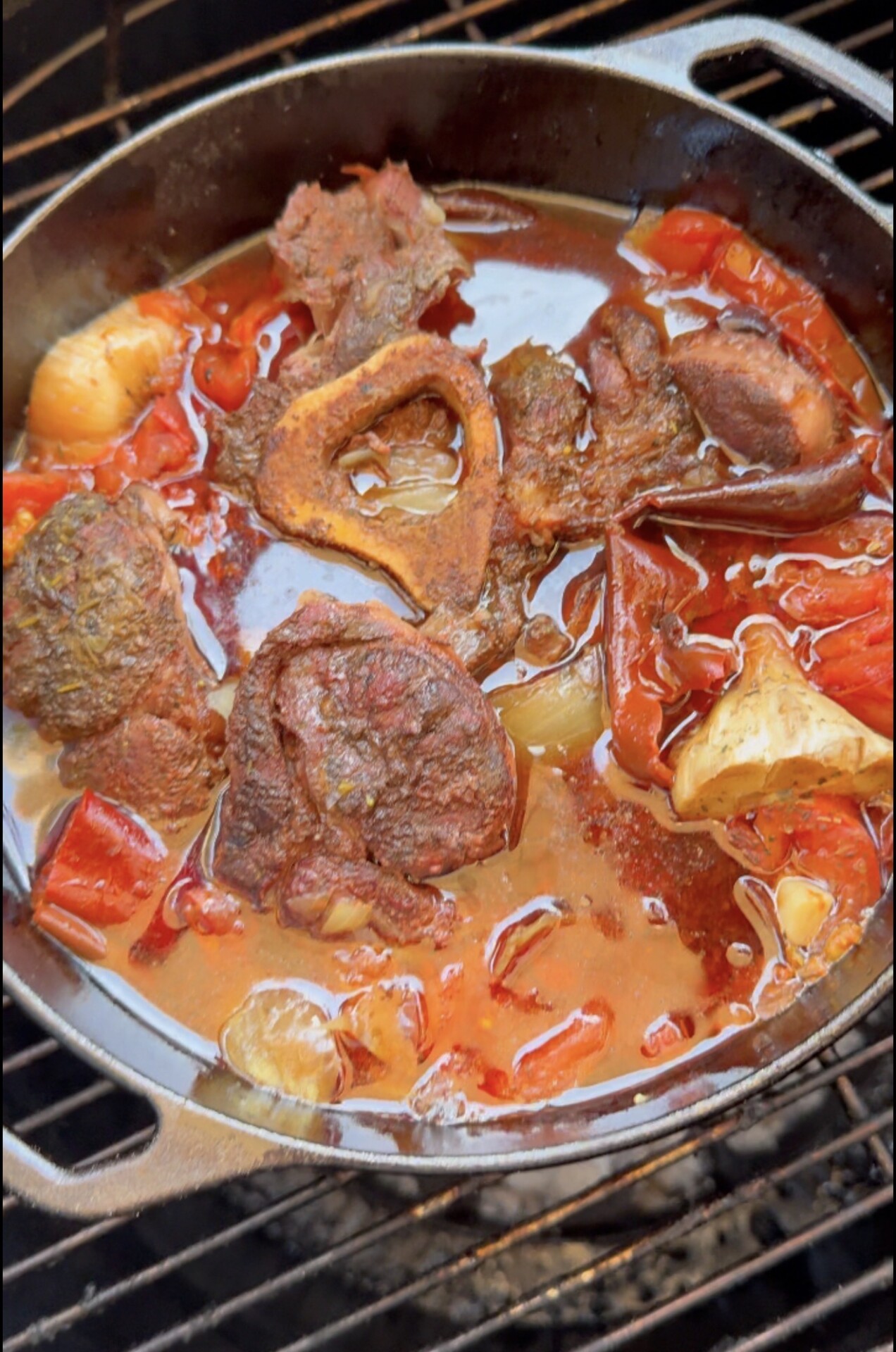 A cast iron pot on a grill contains simmering beef, a bone with marrow, tomatoes, onions, and rich broth, creating a hearty stew.