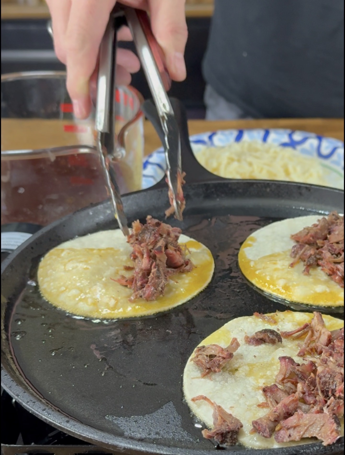 A hand uses tongs to place shredded meat onto corn tortillas frying in a cast iron skillet filled with oil.