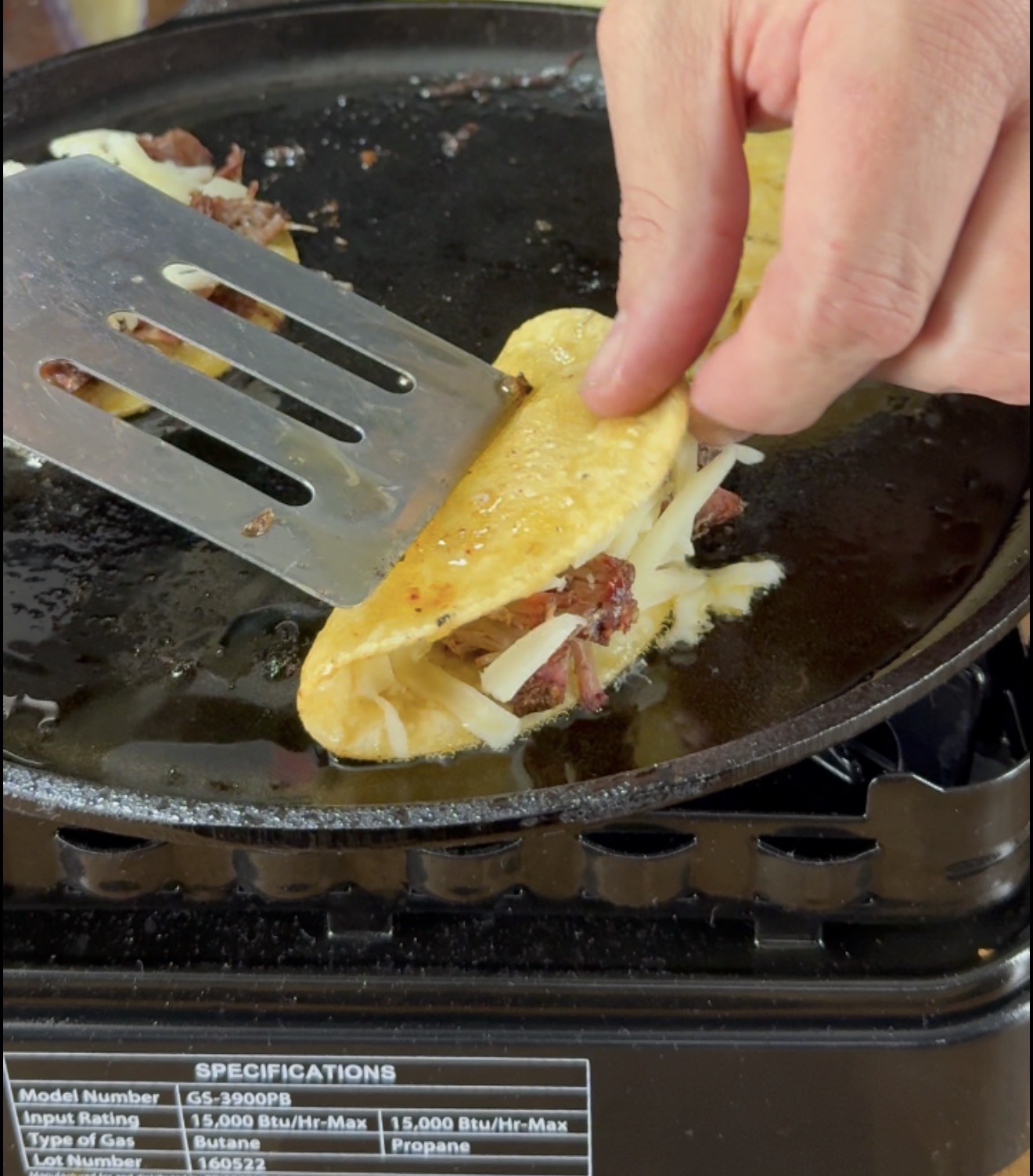 A hand folds a taco with shredded meat and cheese on a hot griddle, using a metal spatula. A label with specifications is visible on the stove.