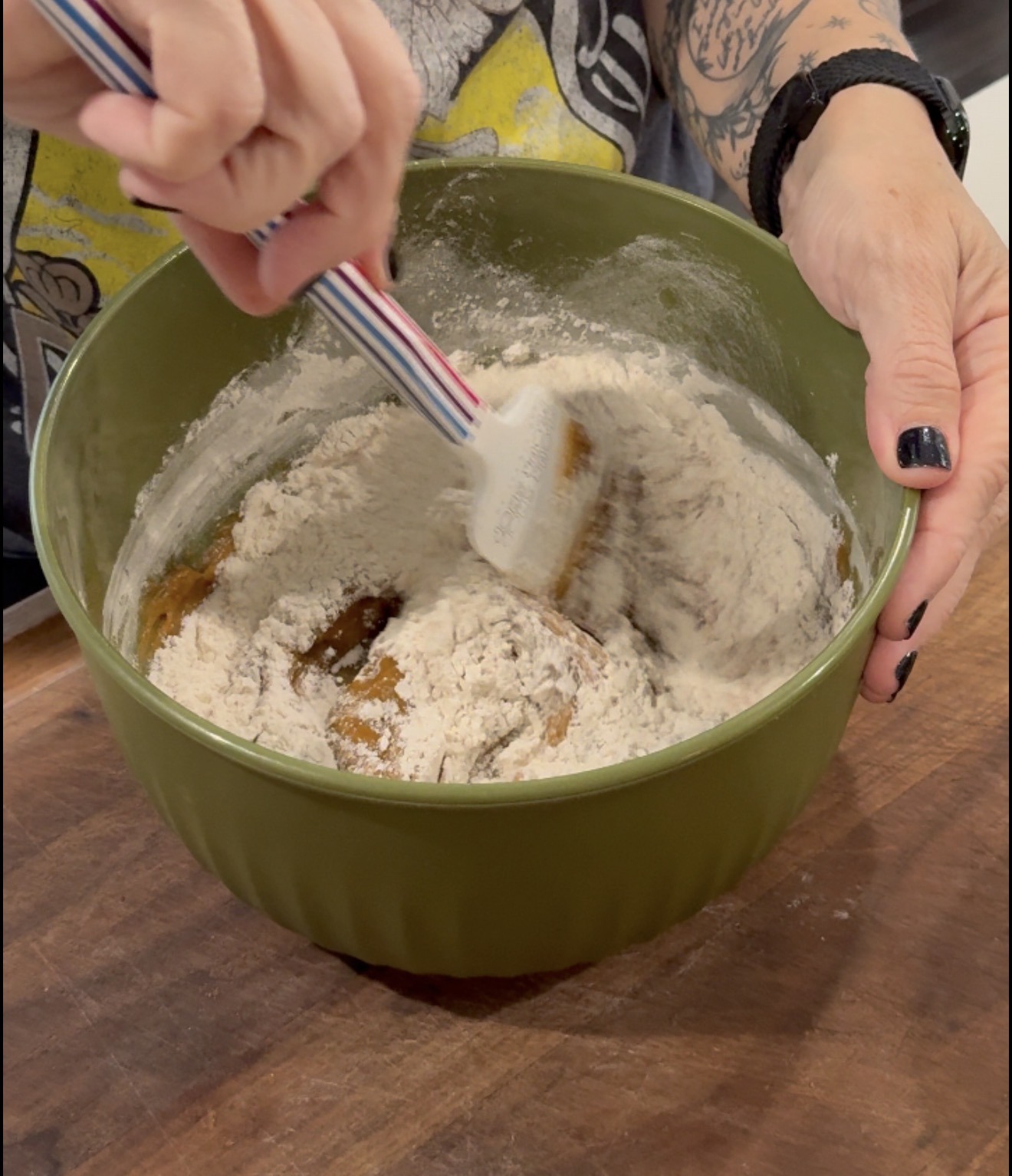 A person with tattoos stirs flour and wet ingredients in a green mixing bowl with a spatula on a wooden surface.