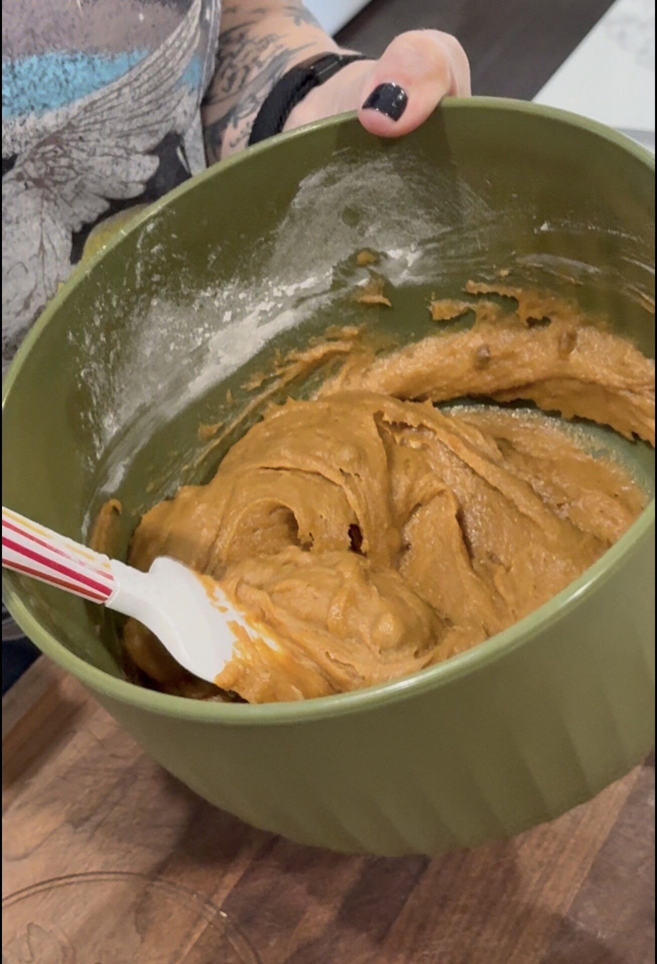 A person holding a green bowl of cookie dough batter and stirring it with a spatula. The person has a tattoo and painted nails.