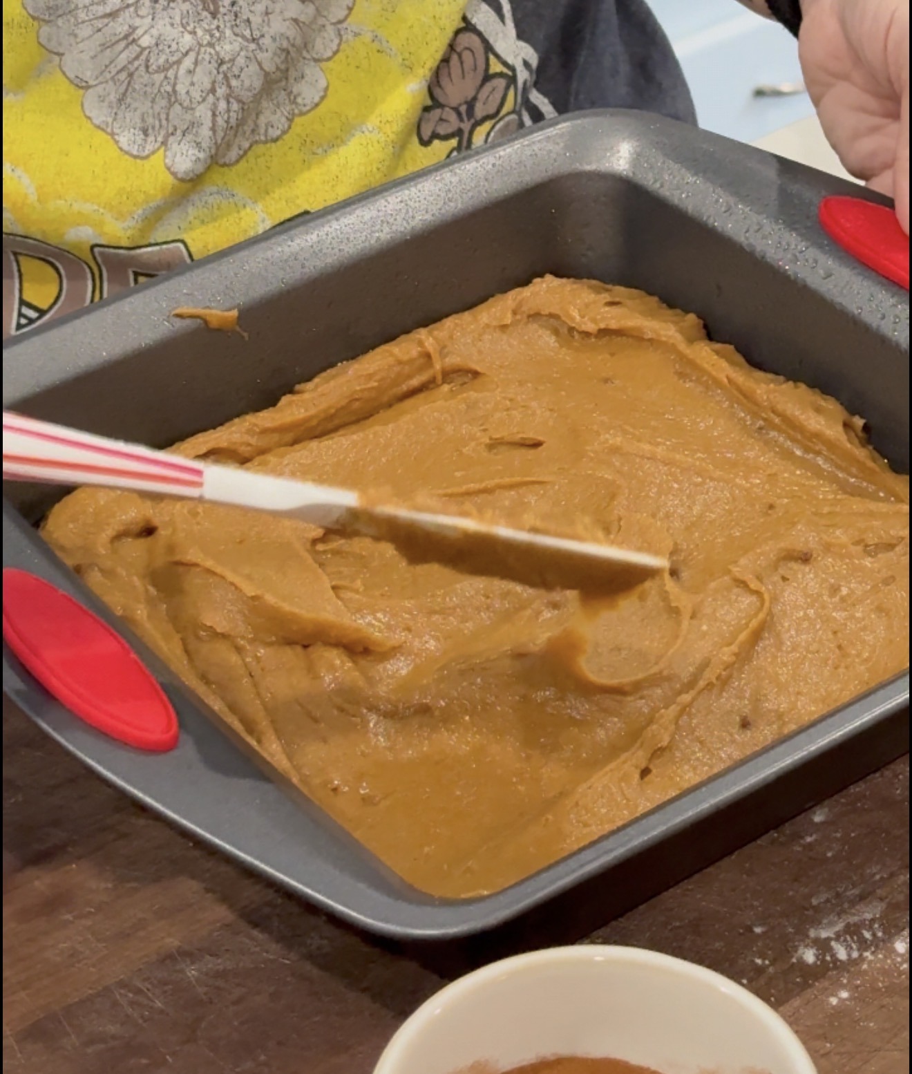 A person spreads a thick brown batter evenly in a square baking pan using a spatula, with a bowl of cocoa powder nearby on a wooden counter.