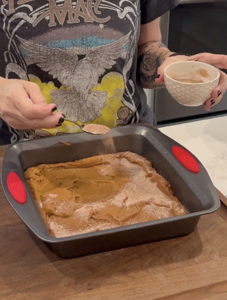A person sprinkles cinnamon sugar from a spoon onto batter in a square baking pan. The person is holding a small bowl and wearing a t-shirt with an eagle design.