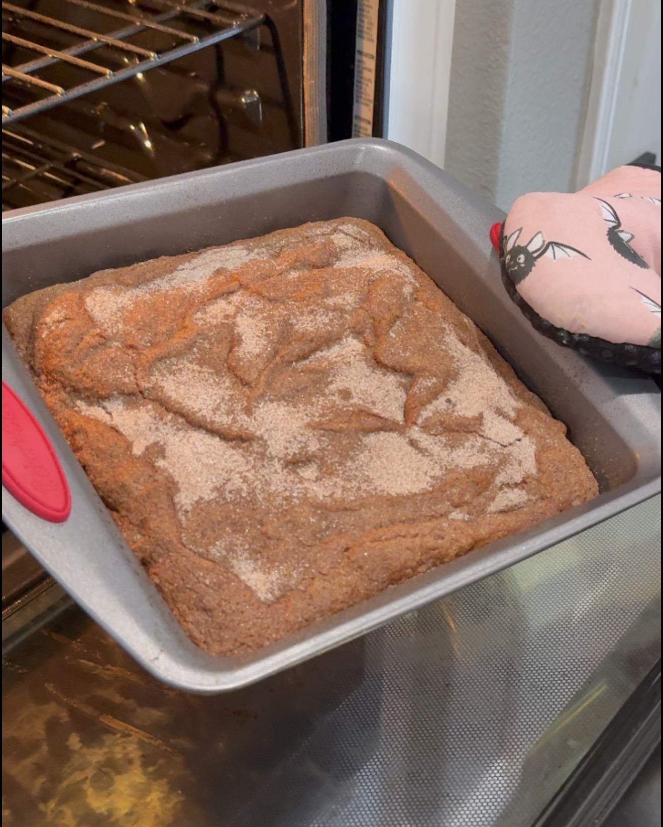 A person wearing a pink oven mitt with a spider design holds a square metal pan of freshly baked cinnamon sugar cake near an open oven.