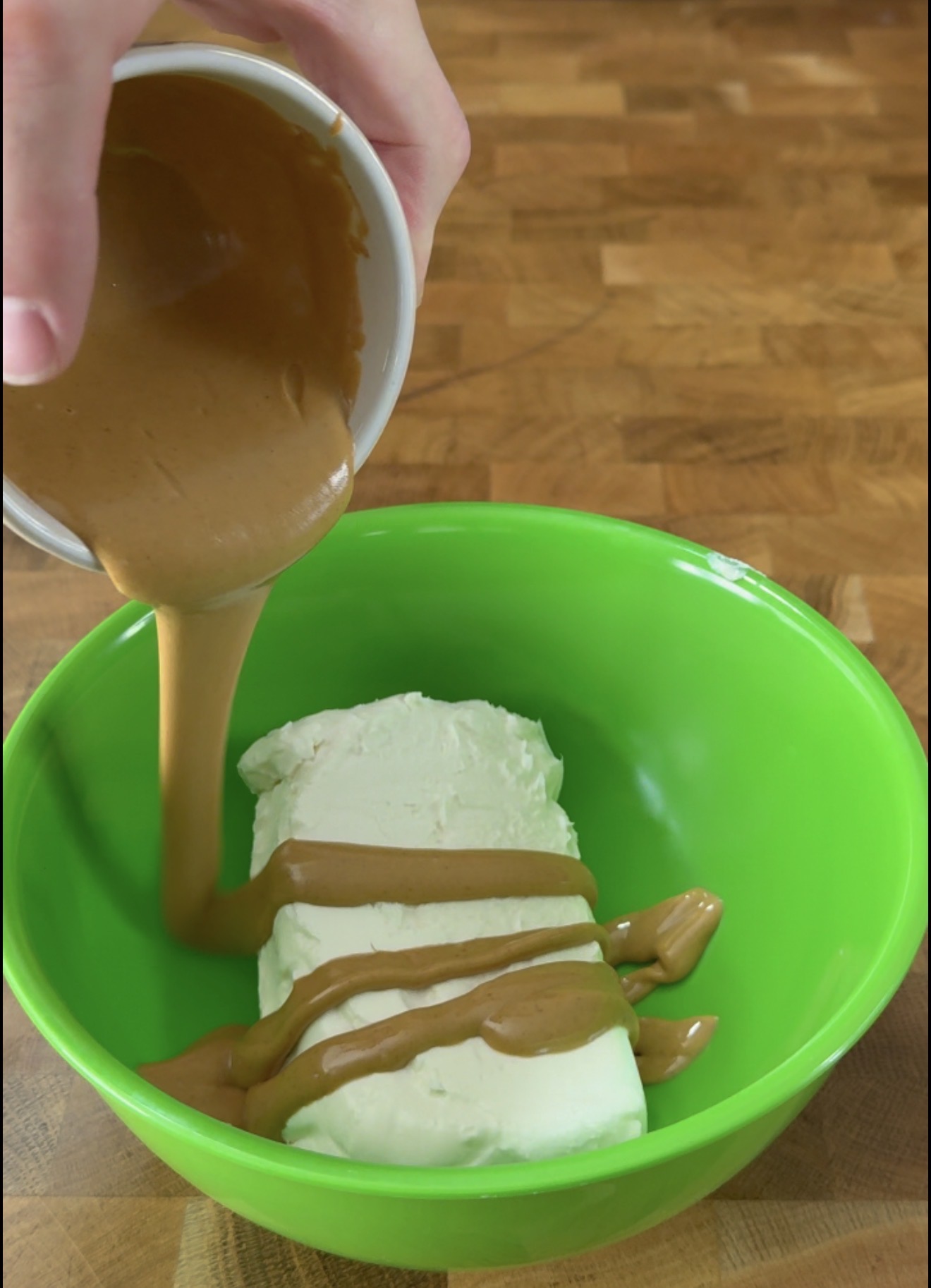 A hand pours creamy brown peanut butter over a block of cream cheese in a bright green mixing bowl on a wooden surface.